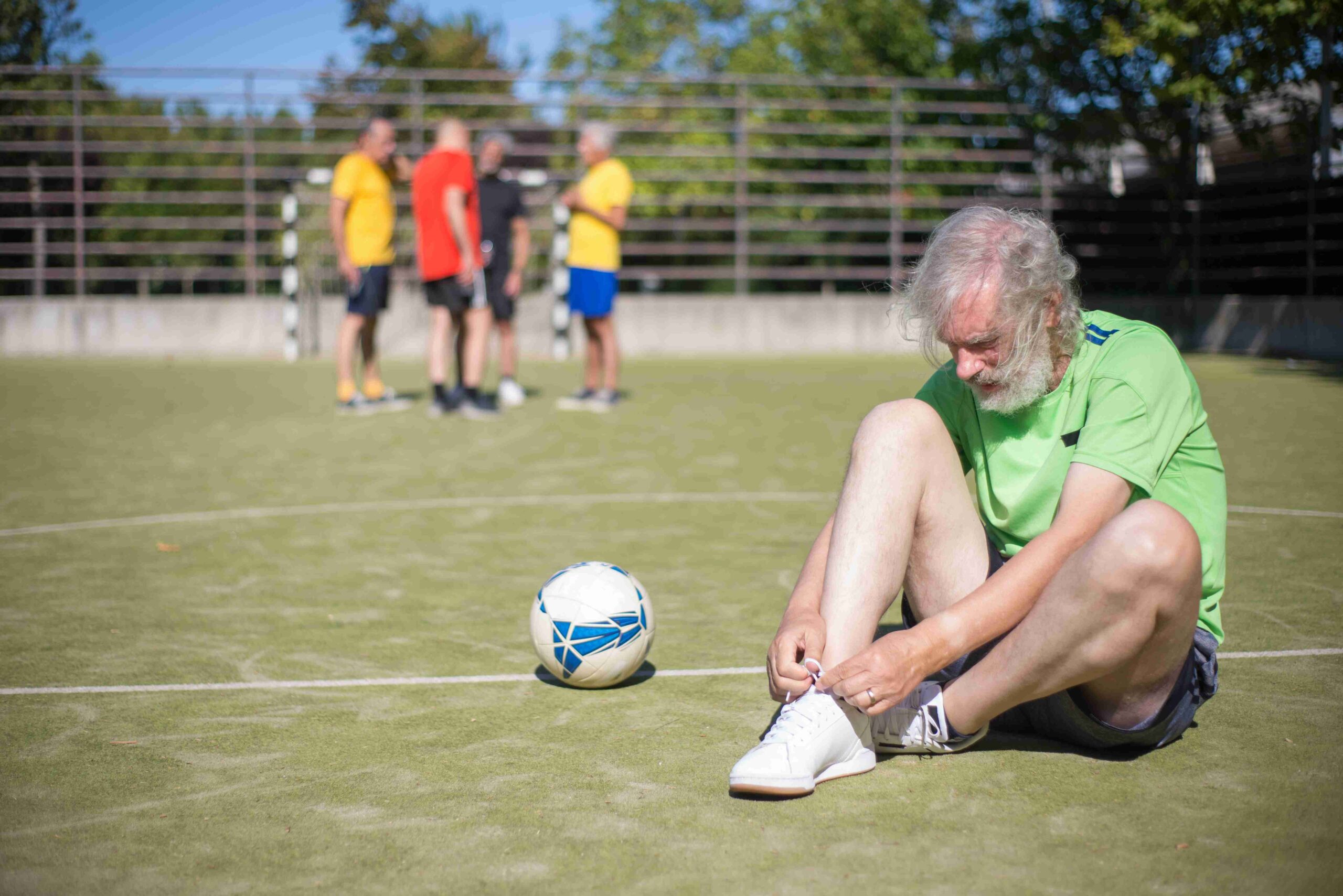 Homem mais velho com camiseta verde e barba branca se dedica a ajustar o cadarço de um tênis ao lado de uma bola de futebol na grama de um campo, enquanto outras pessoas treinam ao fundo.