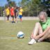 Homem mais velho com camiseta verde e barba branca se dedica a ajustar o cadarço de um tênis ao lado de uma bola de futebol na grama de um campo, enquanto outras pessoas treinam ao fundo.